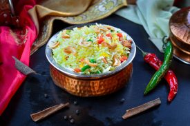 Bright Indian biryani with vegetables and nuts in a copper bowl, garnished with spices and chilies.